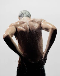 Rear view of a man with wet hair and skin standing under cascading water against a plain white studio background.