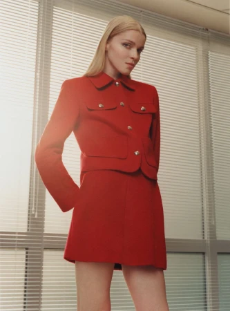 A blonde-haired woman poses indoors wearing a red skirt suit with button details, standing in front of blinds with soft natural lighting.
