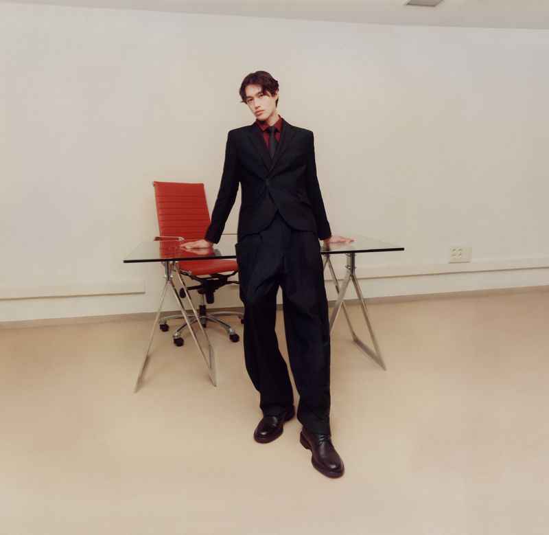 Young adult poses confidently in a dark suit against a white wall, standing near a glass-topped desk and red chair in an indoor setting.