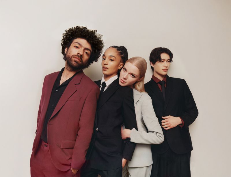 Four young adults in modern business attire pose indoors. Two women and two men look directly at the camera. Studio lit, neutral background.