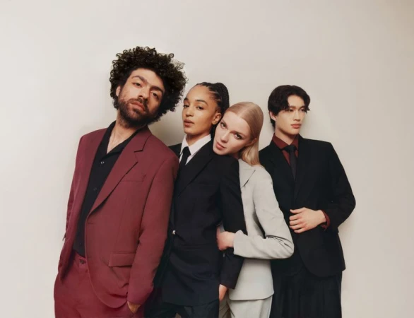 Four young adults in modern business attire pose indoors. Two women and two men look directly at the camera. Studio lit, neutral background.