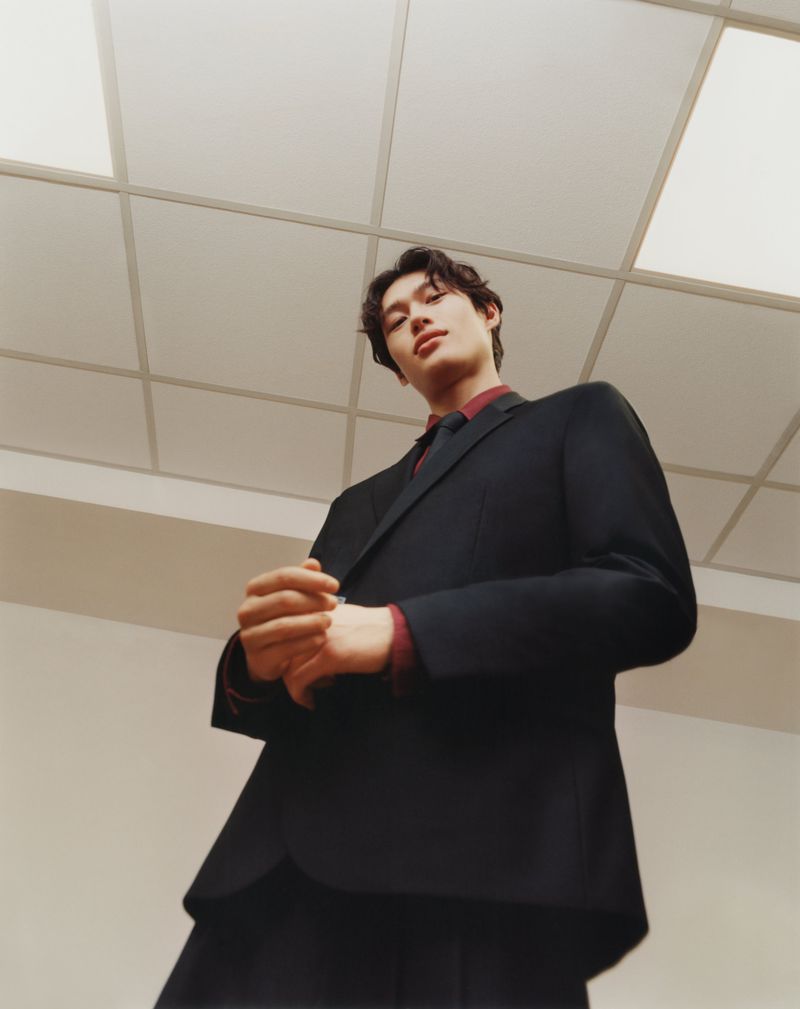Low-angle shot of model Mathieu Simoneau wearing a tailored black blazer and burgundy dress shirt beneath an office drop ceiling.