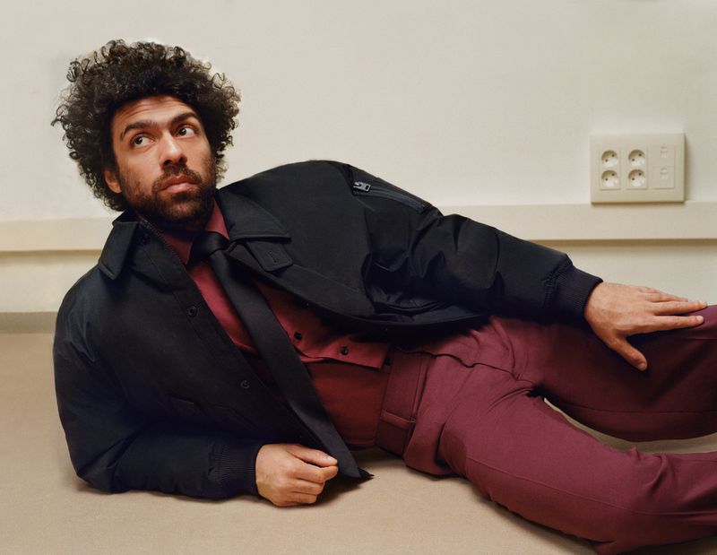 Adult male model poses in a studio, wearing a bomber jacket, red shirt, and trousers, with a neutral background and natural lighting.