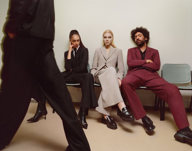 Three models in burgundy, beige, and black suits sit on chairs, while one partially visible model walks in front in black.