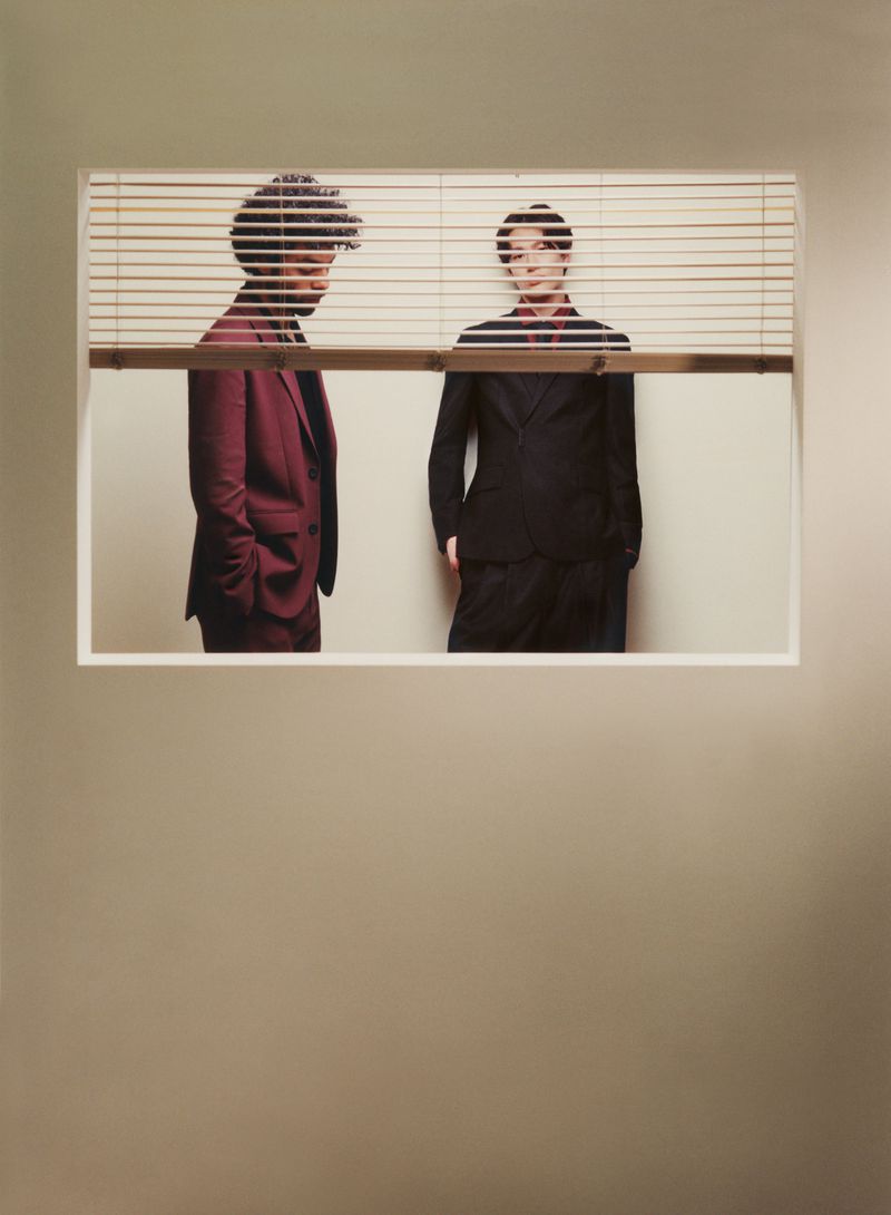 Two stylish men in suits pose behind window blinds, creating a unique visual composition with formal wear and intriguing light play.
