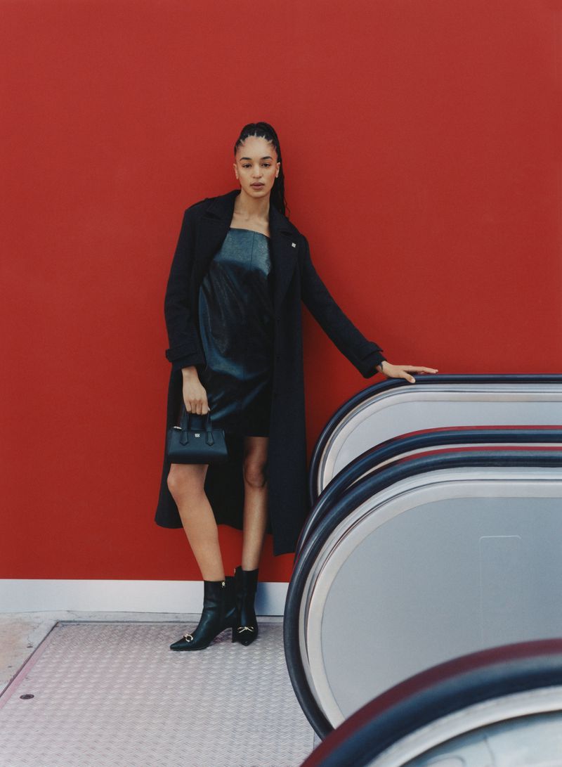 A woman wearing a black coat and dress stands in front of a red wall and next to an escalator, posing for a fashion shot.