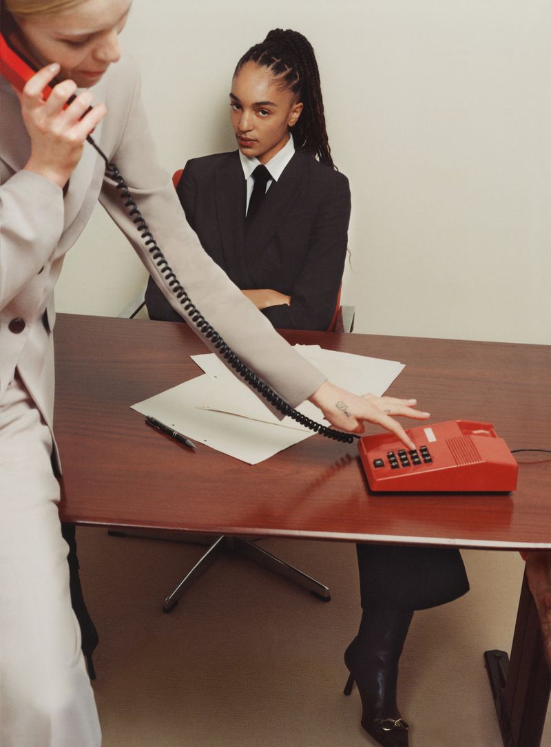 Two businesswomen in suits, one using a red landline phone, in an office setting with a desk and paperwork, under studio lighting.