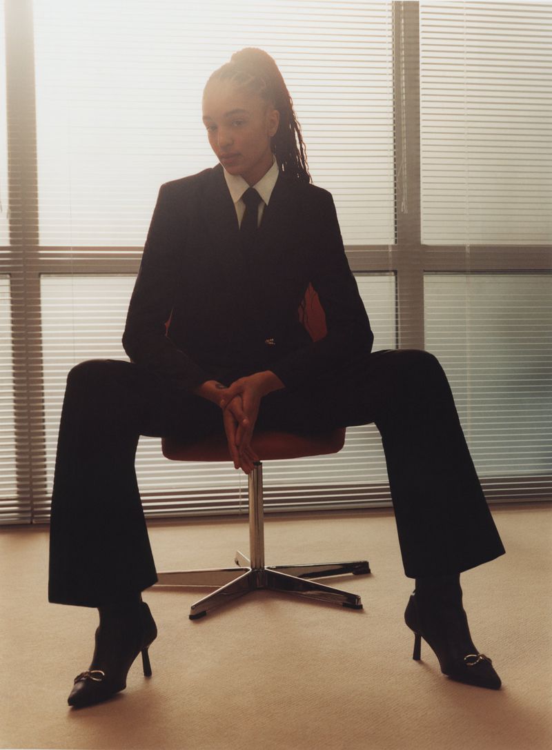 Young woman in dark suit and heels sits on chair in front of window blinds; confident posture, indoor setting with natural light.