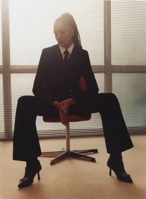 Model wearing a black suit and tie seated on a swivel chair in an office interior with window blinds