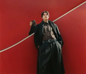 Man wearing a black coat and trousers standing against a vibrant red background with a reflection of a person