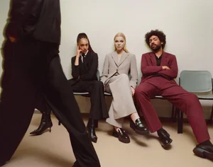 Group of diverse models wearing tailored suits sitting on chairs in an office setting with a figure walking past in the foreground.