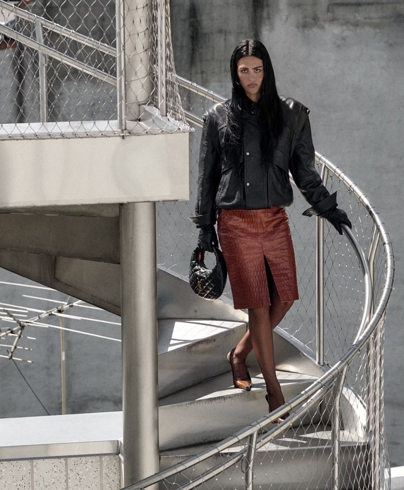 Woman descends a modern spiral staircase wearing a leather jacket, red skirt, gloves, handbag and heels, with concrete wall background.