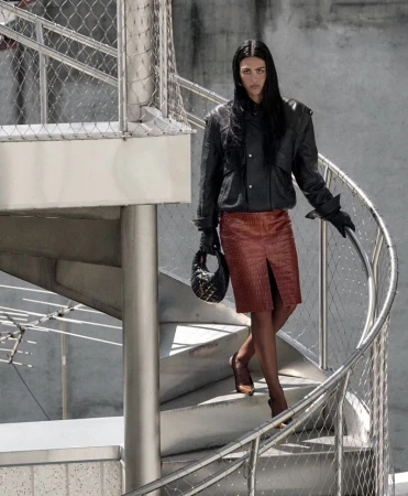 Woman descends a modern spiral staircase wearing a leather jacket, red skirt, gloves, handbag and heels, with concrete wall background.