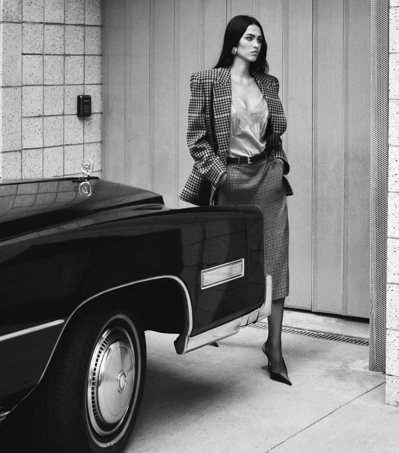 Elegant woman in a blazer and skirt posing next to a classic black car. Classic fashion, monochrome photography in an urban setting.