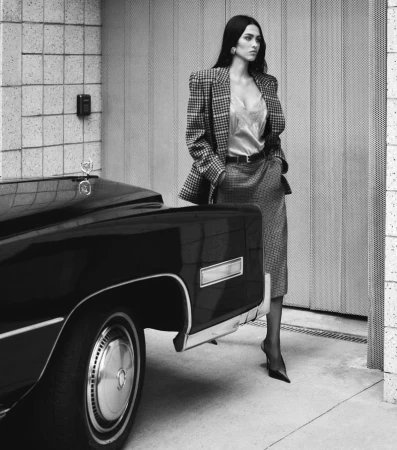 Elegant woman in a blazer and skirt posing next to a classic black car. Classic fashion, monochrome photography in an urban setting.