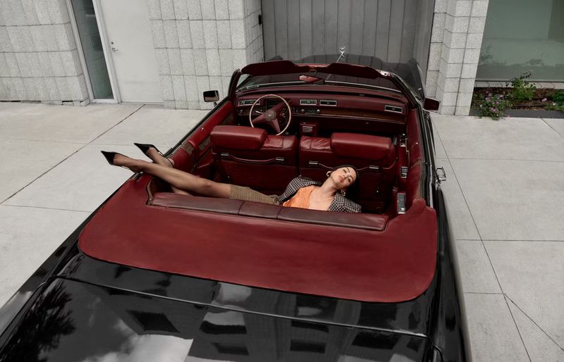 Adult woman poses in a vintage black convertible with dark red interior in front of a modern building with a concrete exterior.