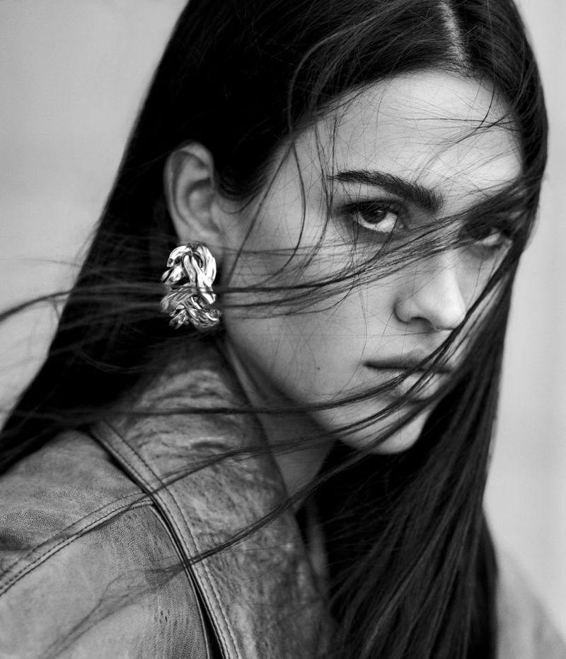 Monochrome portrait of a woman with long dark hair wearing statement earrings and a jacket, looking directly at the camera.