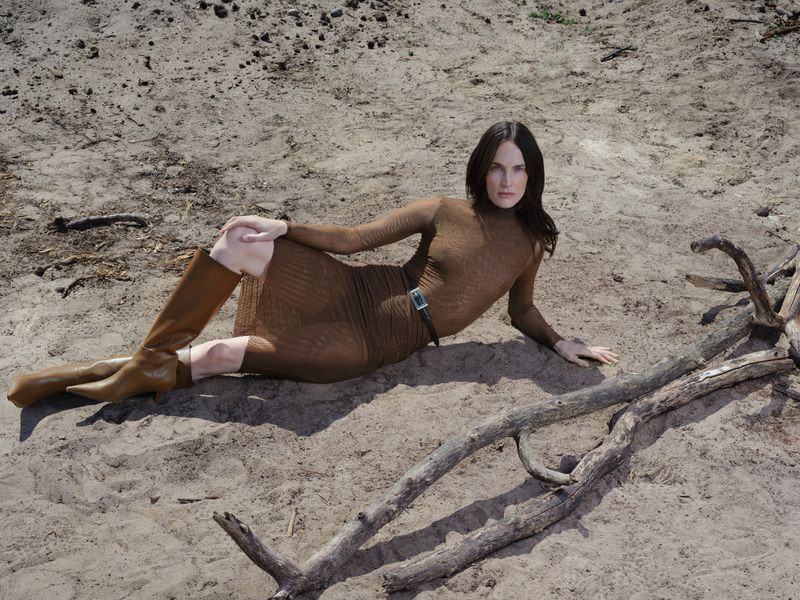Woman in brown dress and boots poses on sandy ground with branches, creating a natural, earthy fashion image with neutral tones.