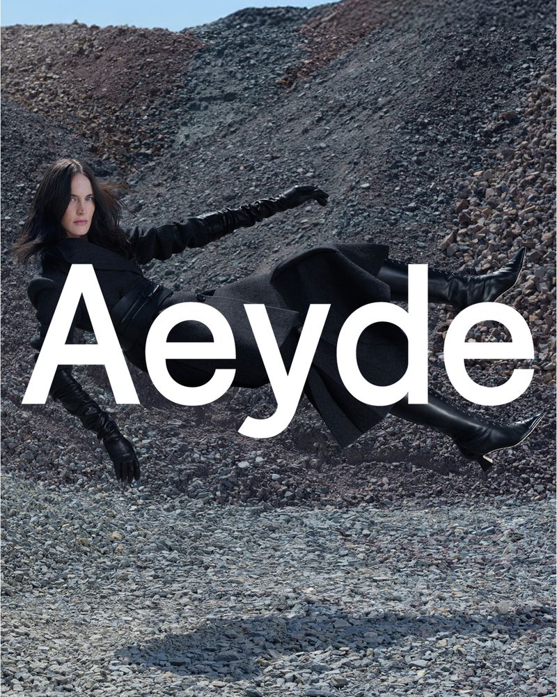 Fashion editorial shot of a woman in a black dress and boots lying on a rocky surface with the word 'Aeyde' superimposed on the image.