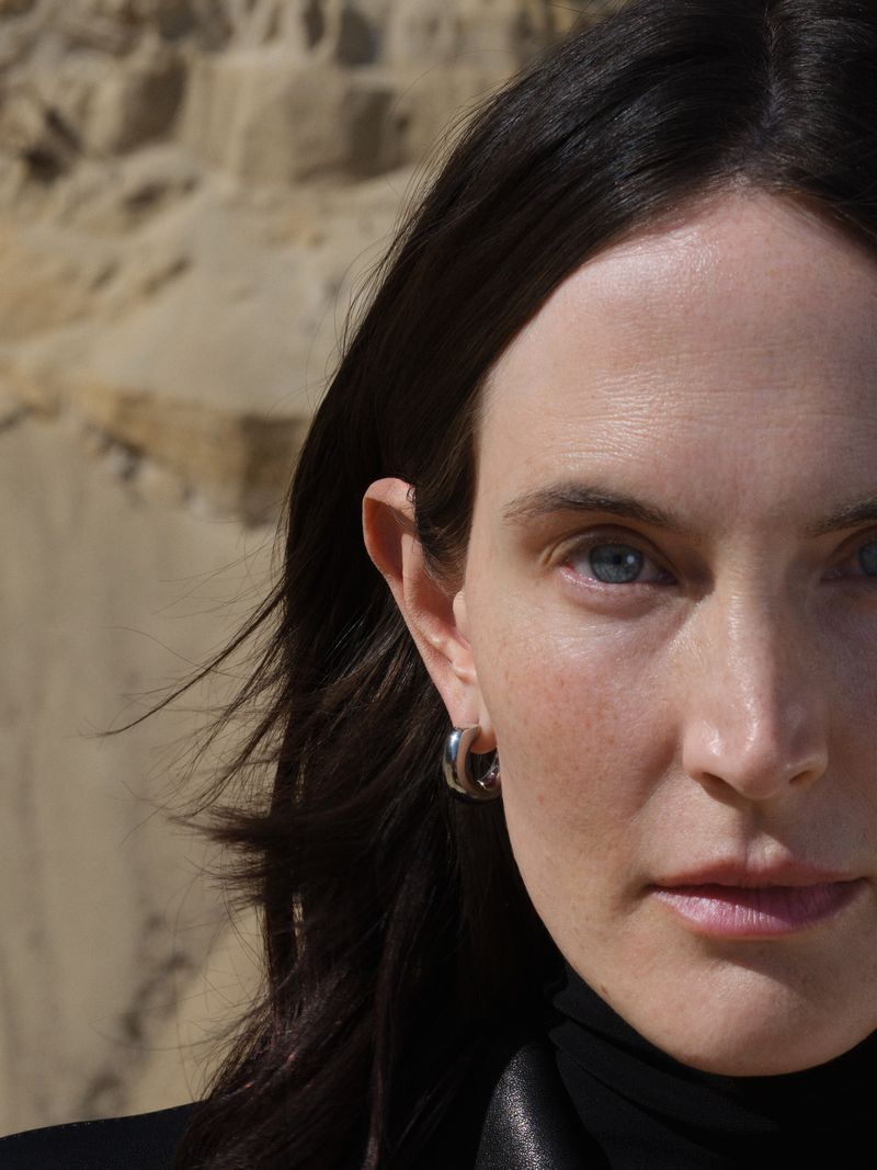 Close-up portrait of a woman with dark hair wearing hoop earrings, set against a textured tan and brown background.