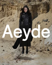Model Jane Moseley in a long black leather coat posing against an eroded sandy cliff for the Aeyde FW25 campaign.