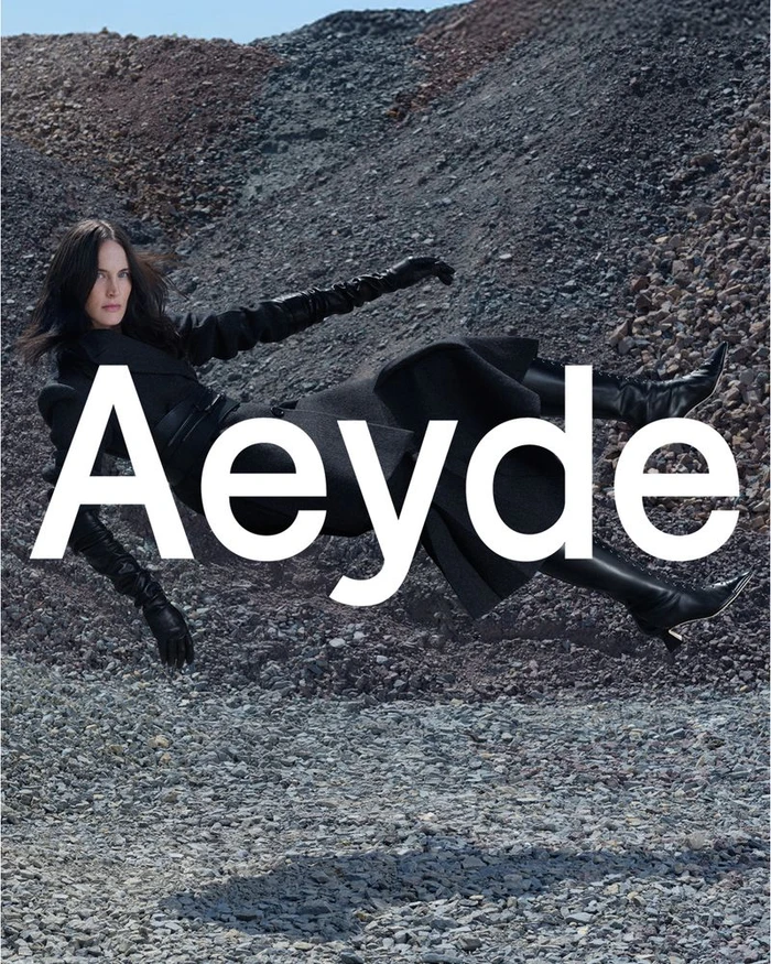 Model Jane Moseley suspended horizontally in mid-air wearing a black coat, long leather gloves, and pointed boots over rocky ground.