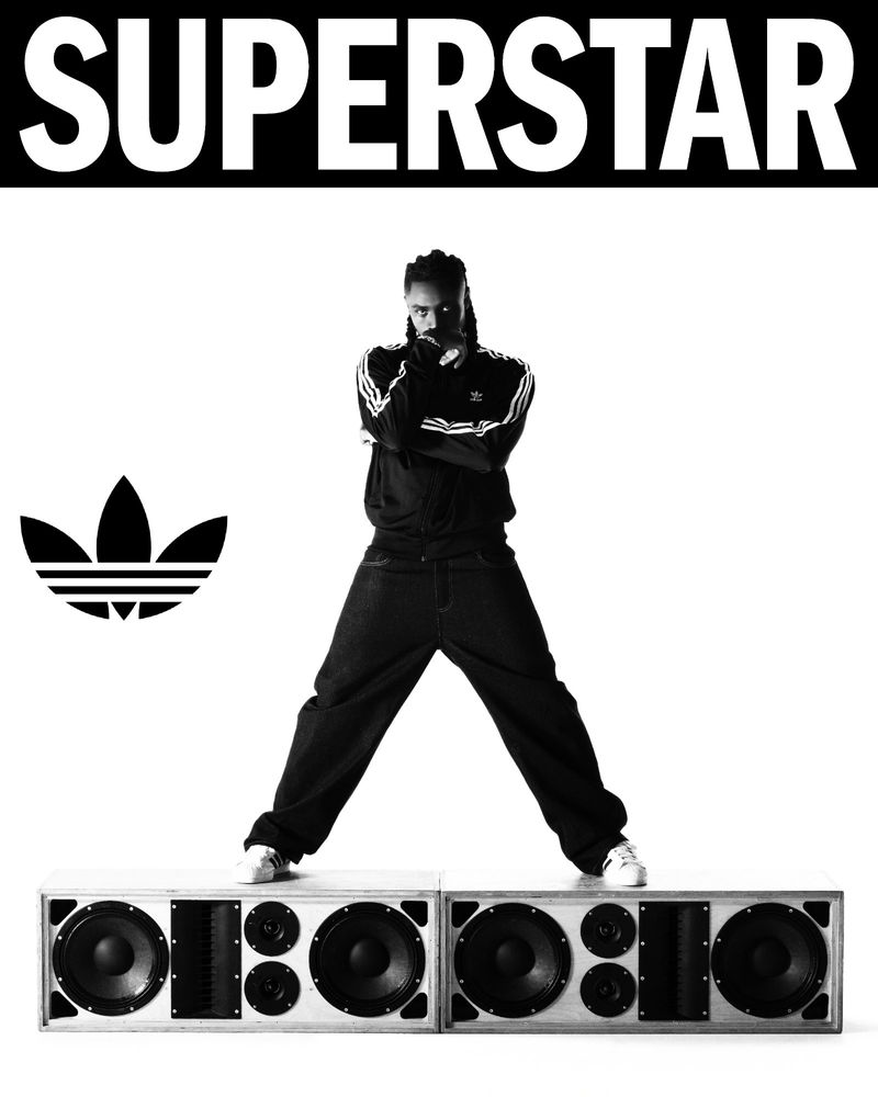 Black and white studio shot of a male in Adidas track suit standing on speaker boxes with the Adidas logo and the word SUPERSTAR above.