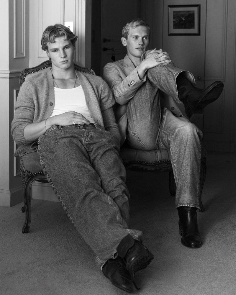 Black and white portrait of two blonde men sitting on an ornate chair. One man wears a cardigan; the other wears a suit jacket.