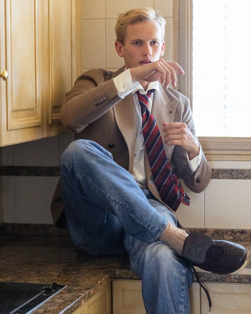 Man in a blazer and jeans sitting on a kitchen counter. He wears a striped tie with brown shoes and socks. Neutral color palette.