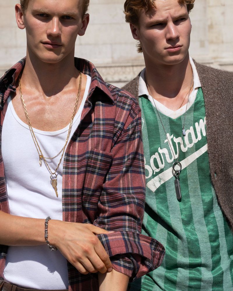 Two young fair-skinned men pose outdoors, one in a plaid shirt and gold chains, the other in a striped tee with a pendant necklace.