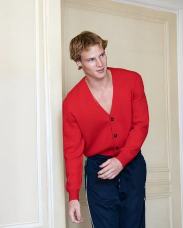 Portrait of a blond male model in a red cardigan and blue track pants leaning against a neutral coloured wall indoors.