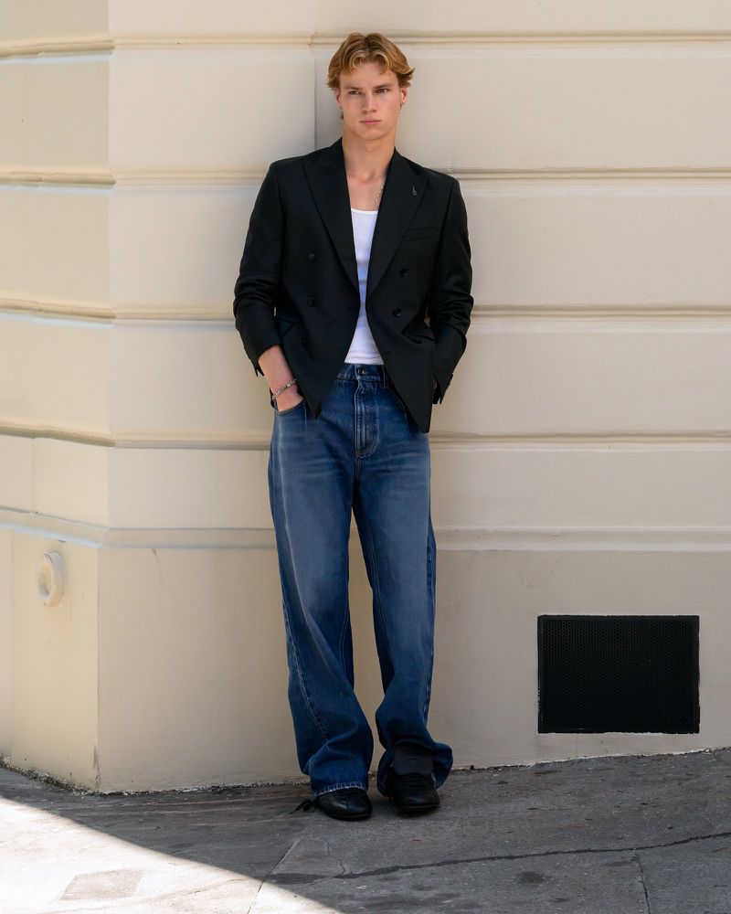 Young man poses in casual-formal attire: dark blazer, white undershirt, blue jeans, and black shoes, standing against a light wall outdoors.
