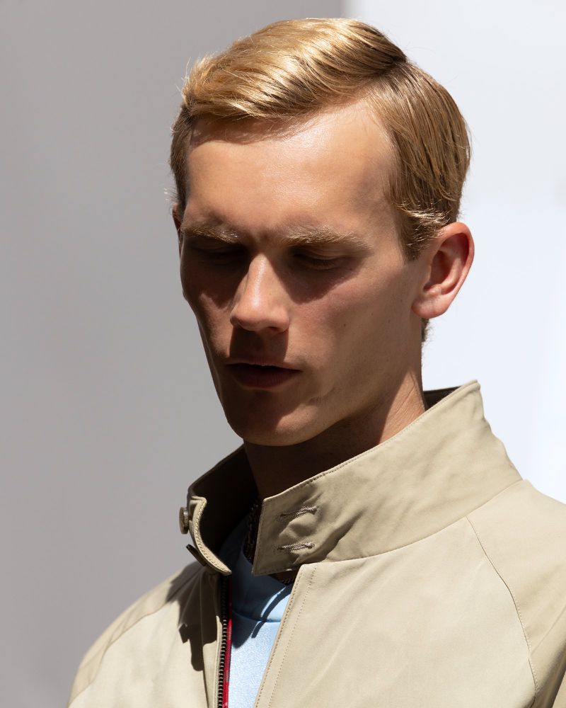 Portrait of a blonde man looking down, wearing a tan jacket against a white background. Head and shoulders shot; natural light.