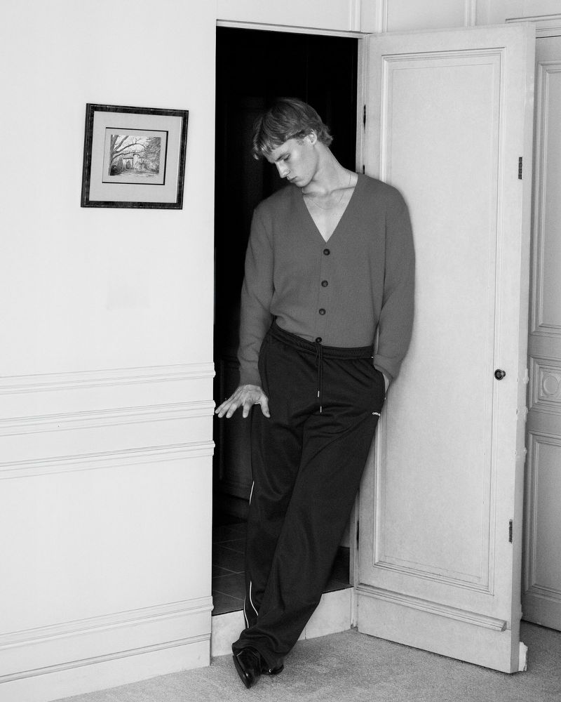 Indoor portrait of a blonde male leaning against a white doorframe wearing a red cardigan and black athletic pants in a minimal interior.