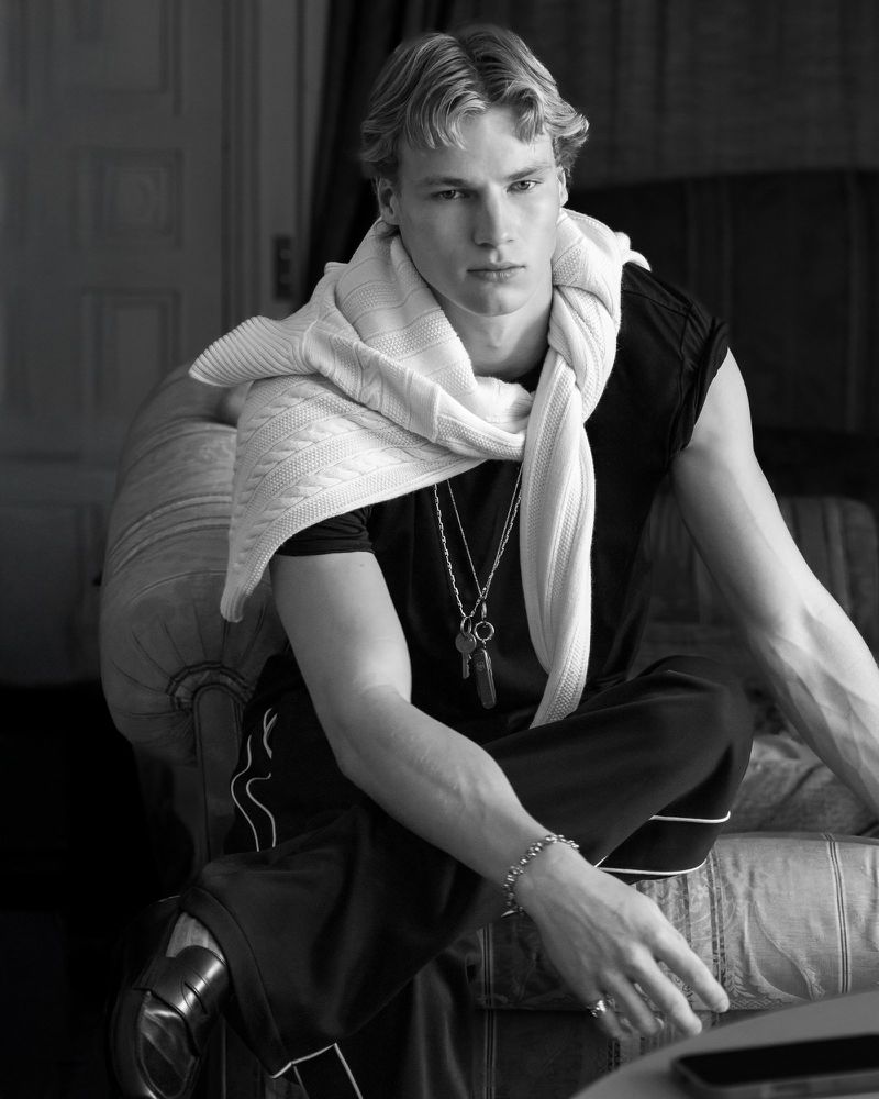 Black and white portrait of a young adult male model with blonde hair wearing a dark shirt, pants, and a light-colored sweater.