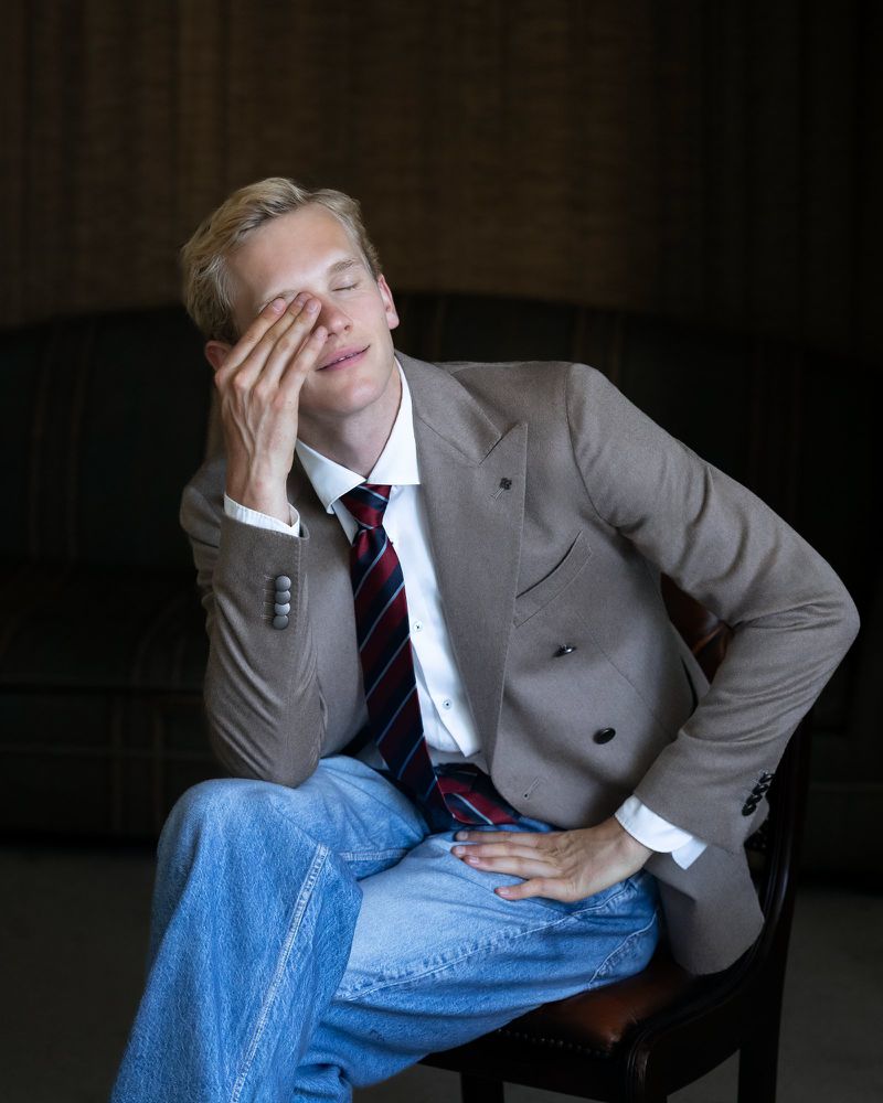 A man in a suit sits with his hand on his face, wearing a white shirt, a tie, and jeans, in an indoor setting against a dark background.