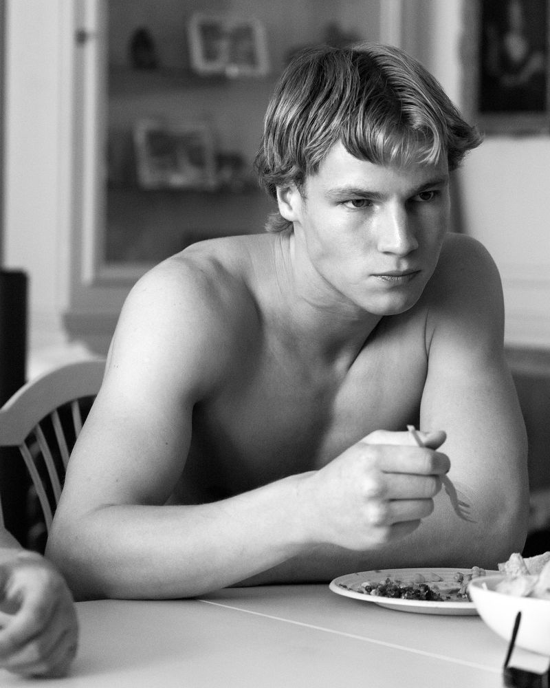 Black and white portrait of shirtless young man seated at a table with a bowl of food, captured with natural light indoors.