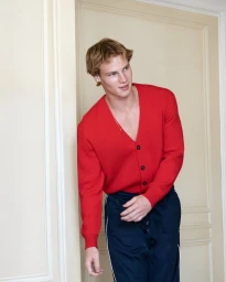 A blond young man wearing a bright red knit cardigan and navy blue track pants with white piping against a cream panelled wall.