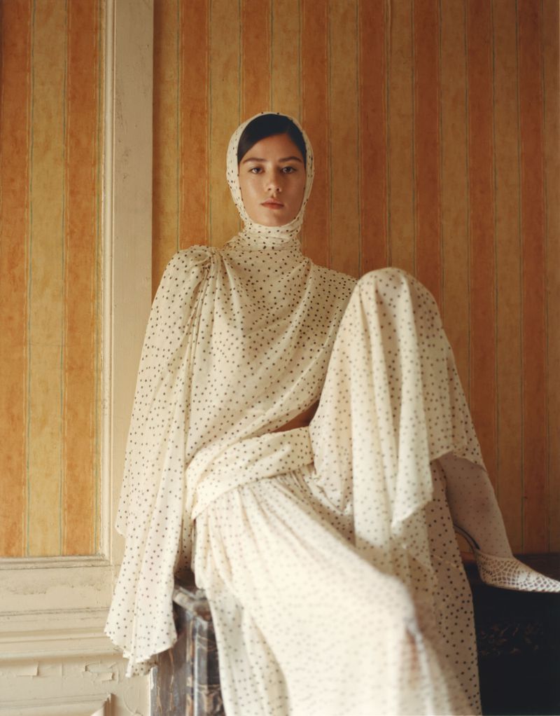 Portrait of a woman in a headscarf and polka dot dress sitting indoors with vertical striped wallpaper behind her in a medium shot.