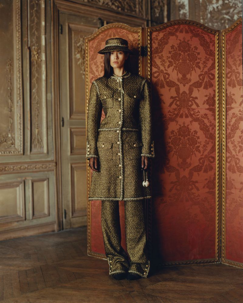 A woman poses indoors wearing a gold tweed suit with pearl trim and a matching hat, standing in front of an ornate screen.