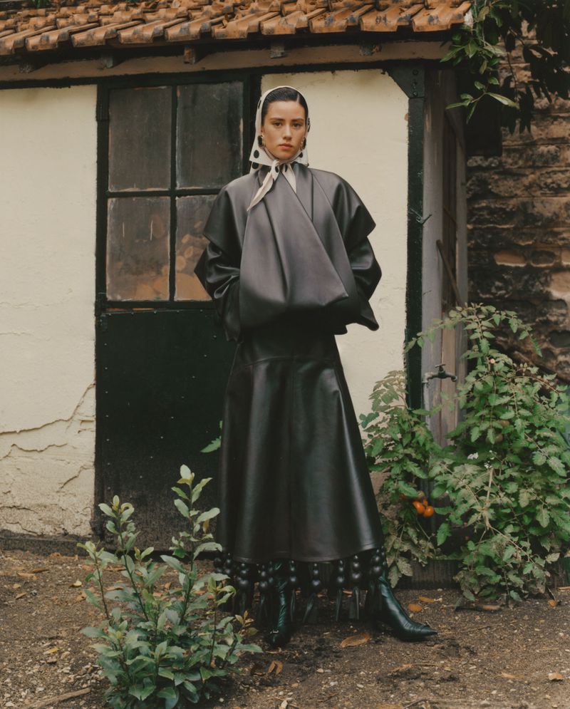 A woman in a dark, oversized leather outfit poses outside a white building with a brown tiled roof, surrounded by green plants.