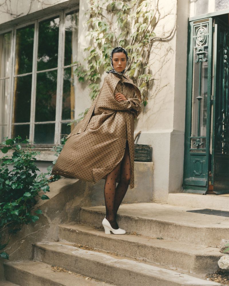 Fashionable adult female in polka dot coat and sheer tights posing on stone steps near building with climbing ivy and green door.