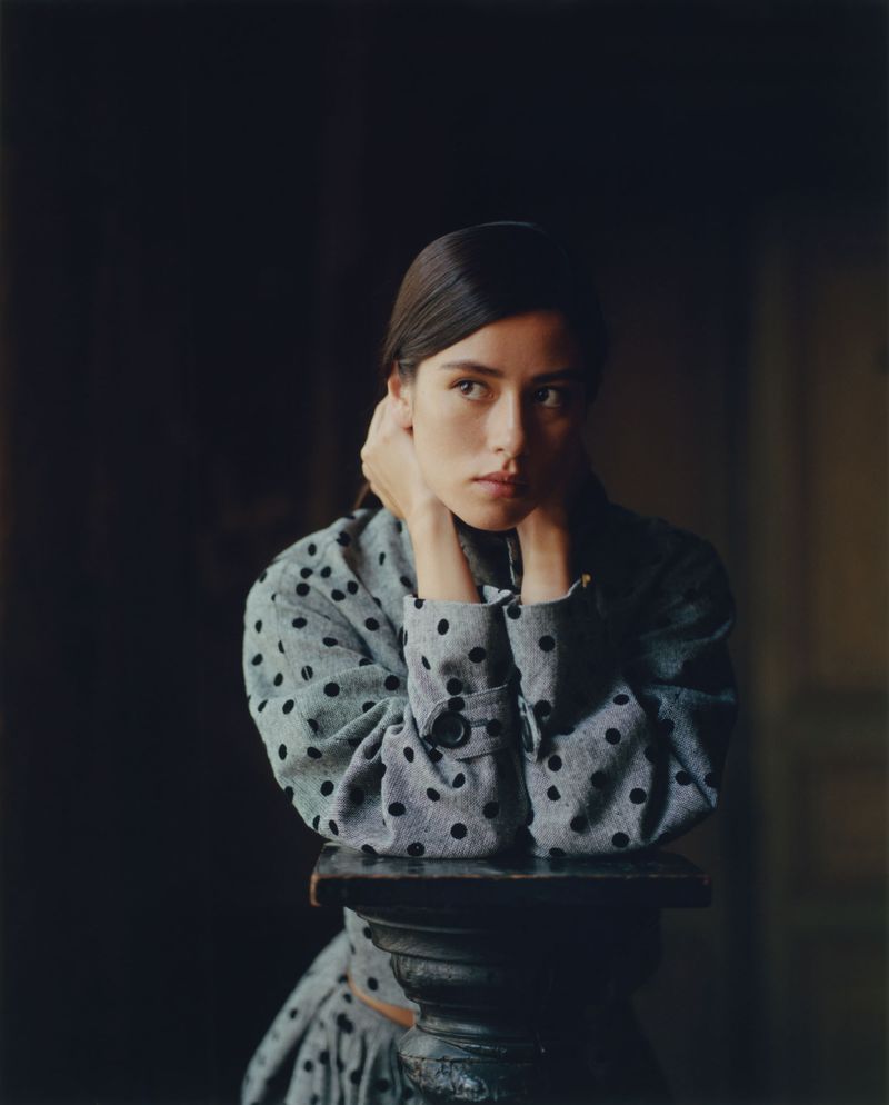 Portrait of a woman in a polka dot top resting her chin on her hands atop a decorative pedestal, set against a dark background.