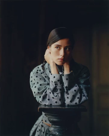 Portrait of a woman in a polka dot top resting her chin on her hands atop a decorative pedestal, set against a dark background.