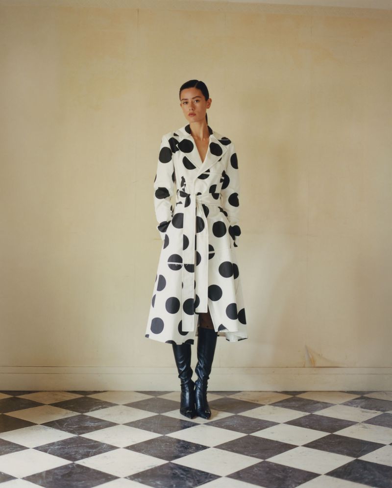 A full-length portrait of a woman in a white, black polka dot dress coat and black boots standing in front of a cream wall and diamond-patterned floor.