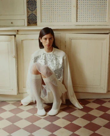 Indoor portrait of a young adult woman in a white top with silver sequins sitting on a tiled floor in front of white cabinets.
