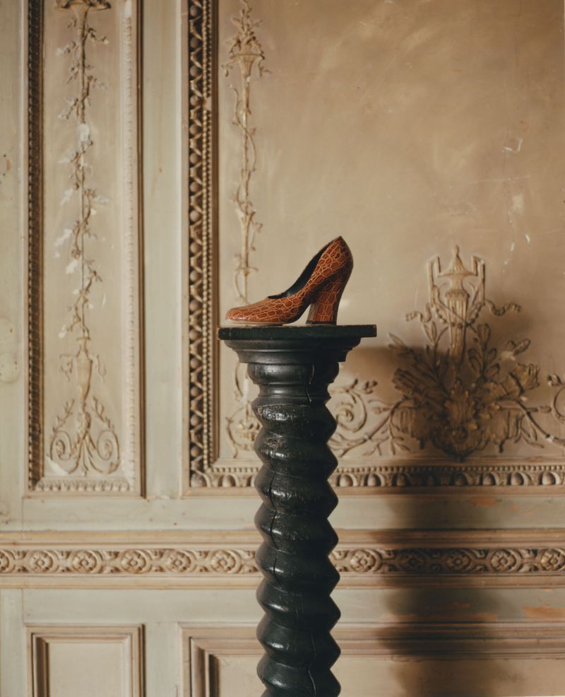 Crocodile-patterned high heel shoe on a black spiral pedestal against a wall with ornate moulding in a classically styled interior.