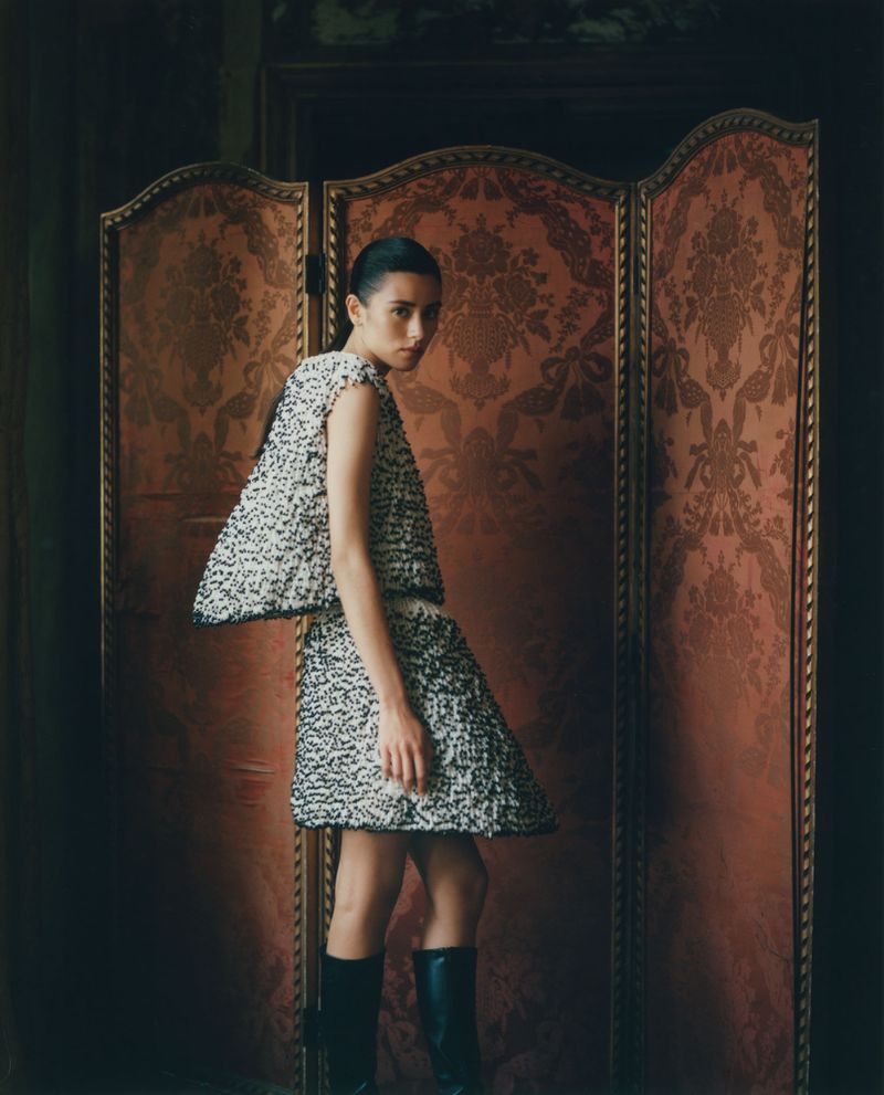 A woman in a textured black and white dress poses in front of a decorative gold screen in a fashion editorial style.