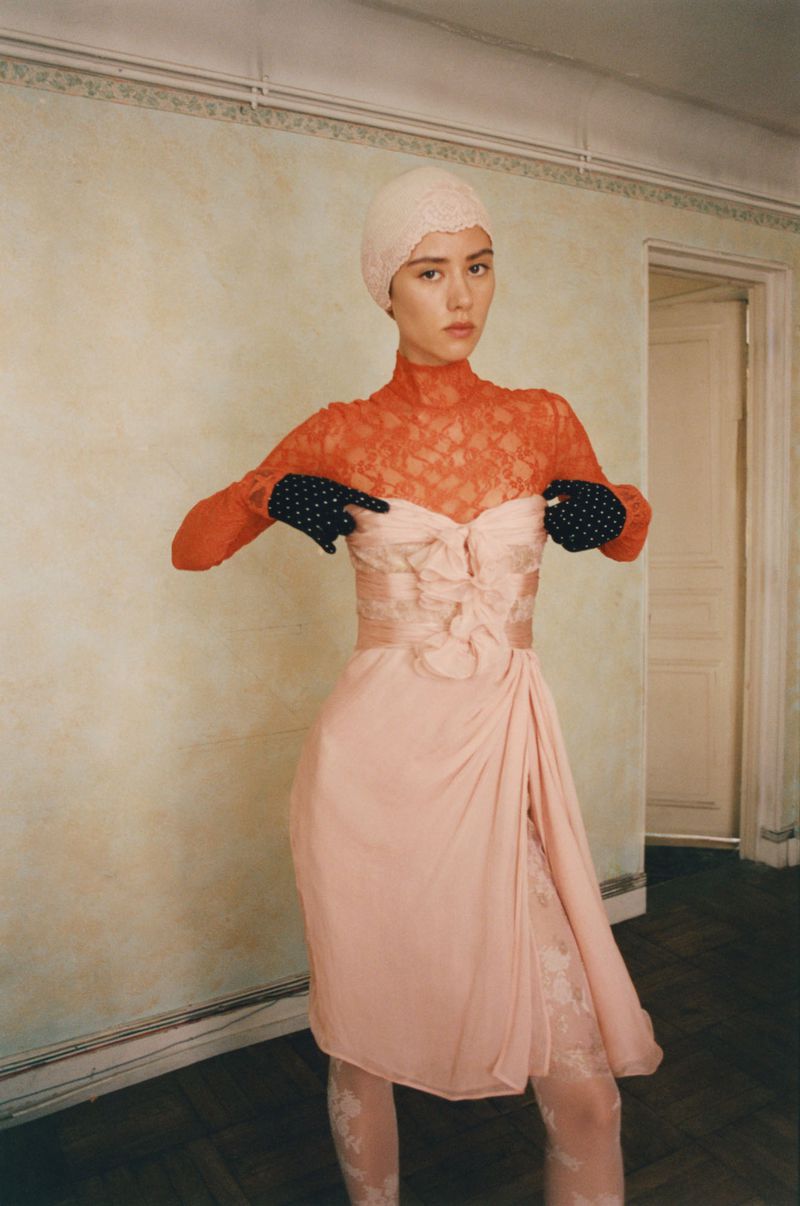 Fashion shot of a woman in a peach-colored dress, lace top, and gloves posing in an indoor environment with neutral-colored walls and a door.