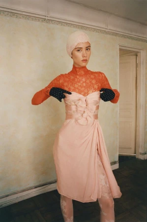 Fashion shot of a woman in a peach-colored dress, lace top, and gloves posing in an indoor environment with neutral-colored walls and a door.